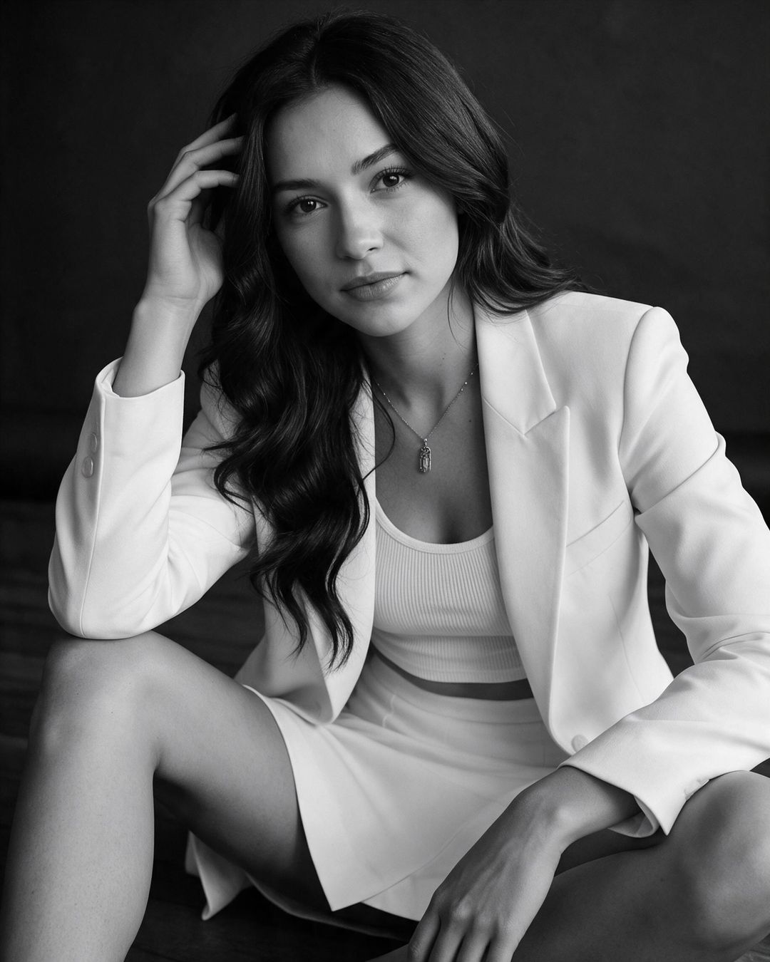 Monochromatyczny portret kobiety w białym garniturze siedzącej na podłodze przy ciemnym tle, efekt sesji AI za darmo.