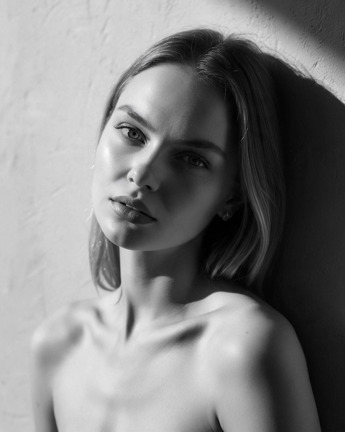 Portret kobiety z odsłoniętymi plecami przy betonowej ścianie, mocne światło i wyraziste cienie.